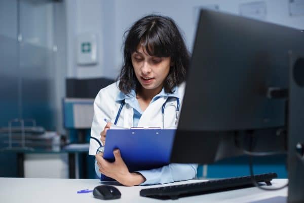 A practitioner wearing a lab coat takes notes in front of her computer.